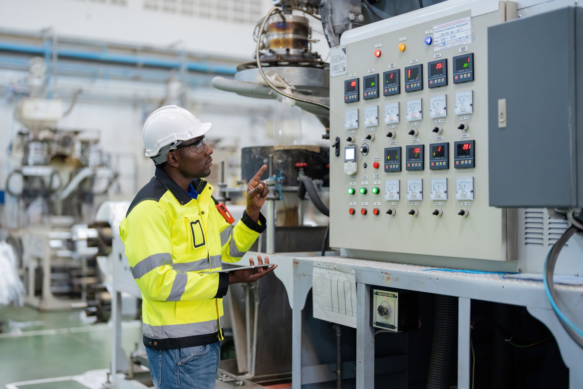 American engineer holding a tablet and pointing his finger at the circuit board controlling the production circuit of a machine in the plastic and steel industry Wear uniforms and helmets.